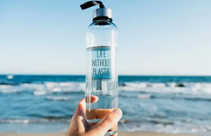 closeup of a caucasian man holding a glass reusable water bottle with the text life without plastic written in it, on the beach, with the ocean in the background; Shutterstock ID 1338555476; Purpose: Plastic free travel tips; Brand (KAYAK, Momondo, Any): momondo